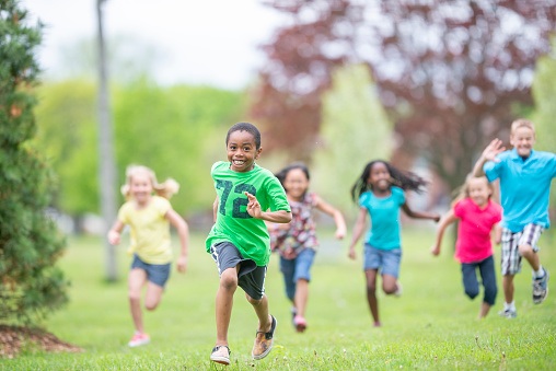 A large group of school aged children run through the grass together during day camp.  They are each dressed casually in colorful t-shirts as they smile and enjoy the summer day.