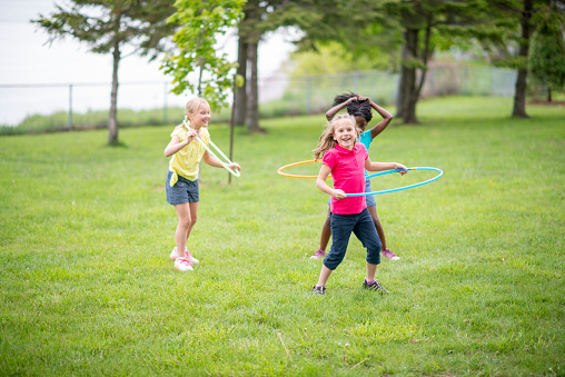 A small group of school aged girls stand in the grass as they Hula-Hoop together.  They are each dressed casually in shorts and t-shirts as they play during day camp.