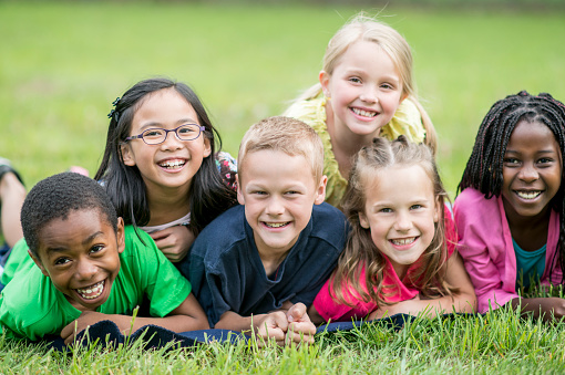 A small group of school aged children lay in the grass, stacked one on top of the other as they pose for a portrait.  They are each dressed casually in colorful t-shirts as they spend time together during day camp.