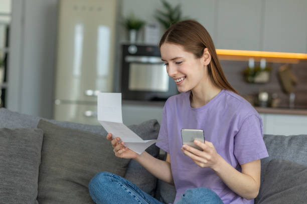 Happy young woman using mobile app to paying household bill, taxes or insurance use secure internet banking. Female holding bank letter reading good news, closed her bank loan stock photo