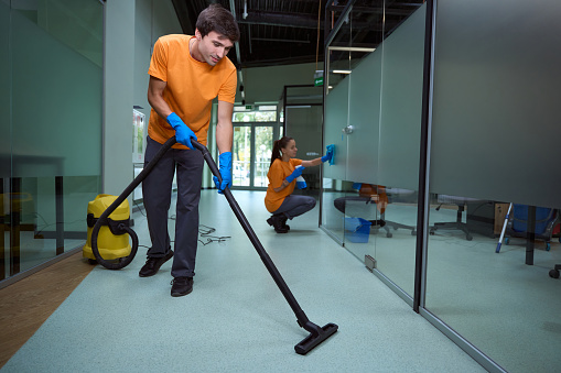 Young man vacuuming the floor while his collegue cleaning the glass surface in the building