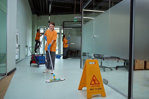 Careful female cleaner using wet mop to wash the floor beside the yellow caution sign