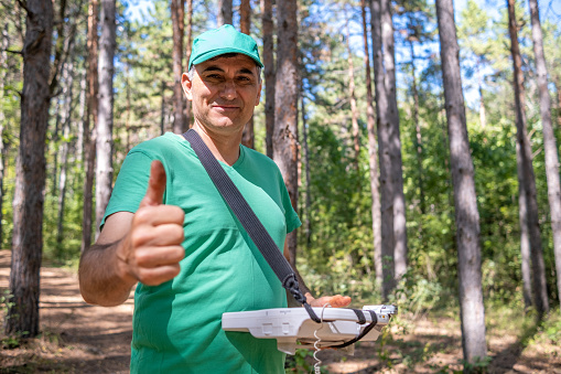 A map maker in a green t-shirt, green baseball cap, and thumbs up in the woods with a specialized computer and GPS. The focus is on the face.