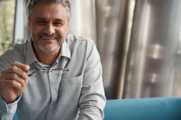 istock Friendly gray-haired businessman sits on sofa,holds glasses in his hands 1436525269