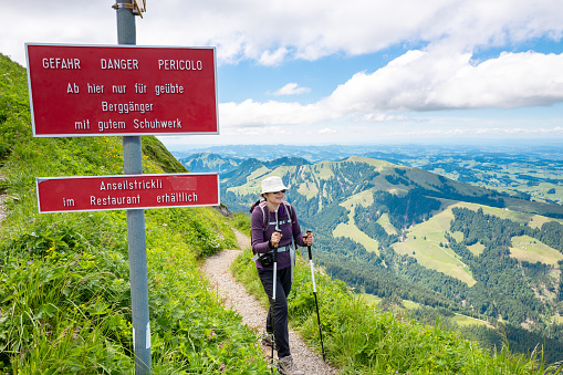 Senior Asian woman hiking the Swiss Alps - warning sign for hikers.  sign: From here only for experienced mountaineers  with good footwear. Rope available at restaurant.
