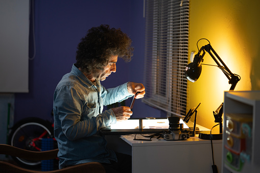Photo of mature adult man sitting on desk and examining 35mm film negatives on light box. a desk lamp is seen on desk. Wall is yellow. He has long curly hair. Shot indoor with a full frame mirrorless camera.