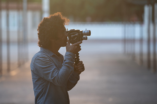 Photo of adult man filming via analog old fashioned antique home video camera. Selective on model. He has curly hair and wearing a jean shirt. Shot in outdoor with a full frame mirrorless camera.