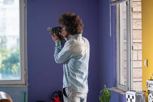 Photo of adult man filming via old fashioned analog home video camera. He is standing in front of a purple colored indoor wall. Shot with a full frame mirrorless camera.