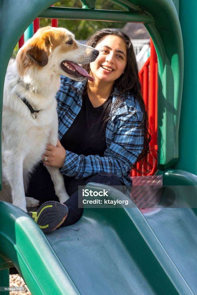 Indian young adult enjoys some fun time with her dog at local dog park and playground Young woman holds onto her Great Pyrenees dog that playfully went to the top of the playground slide Diversity Stock Photo