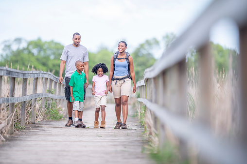 A small African family of four hike across a boardwalk together while on vacation.  They are each dressed comfortably in casual clothing and the mother is carrying a backpack with supplies for their journey.