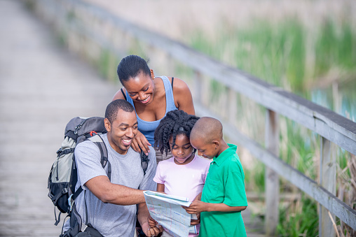 A small African family of four lean in together to look at a map as they navigate their hike.  They are each dressed casually in t-shirts and shorts as they enjoy a warm summers day.  The father is carrying a large backpack of supplies for the family.