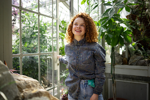 Curly ginger young lady cheerful smiling while walking through greenhouse, wearing denim