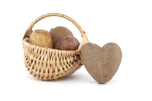 Heart shaped potato next to a wicker basket with potatoes of various types and colors isolated on white background