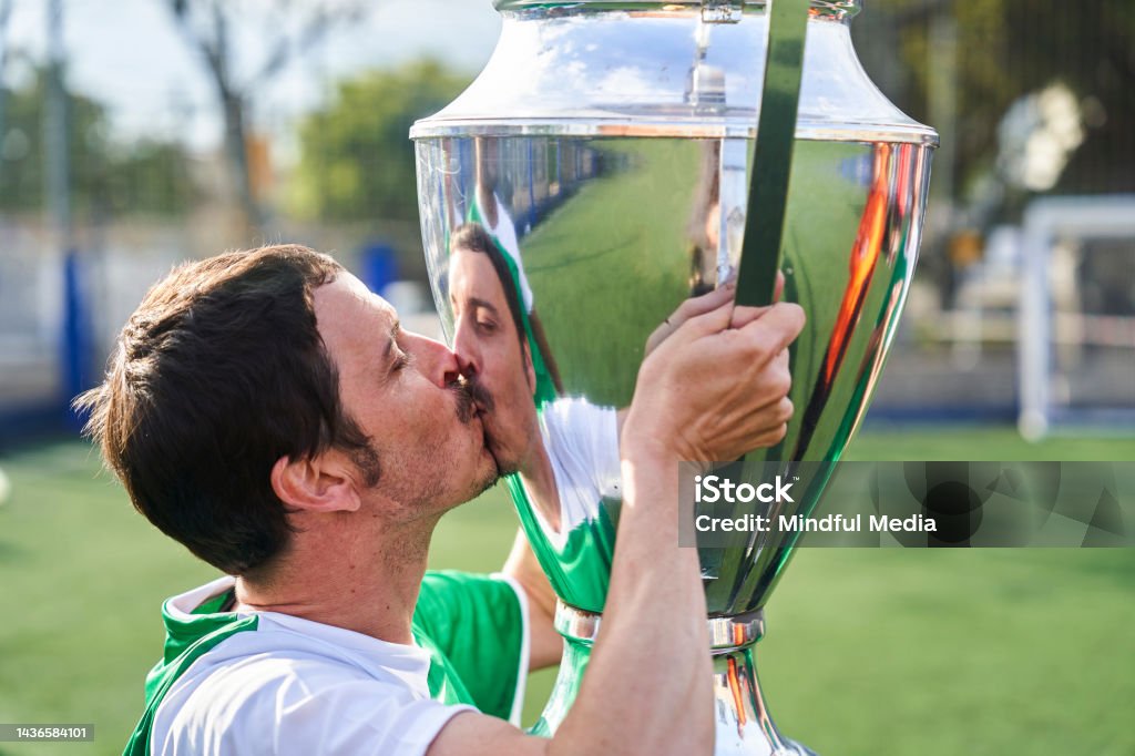 Male amateur soccer player kissing big trophy cup after winning tournament Mid-shot of male amateur soccer player kissing big trophy cup after winning local tournament Argentina Stock Photo
