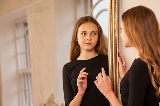 Portrait teenager girl 12-13 year old with long hair, posing at mirror, looking reflection fashion model in stylish black elegant dress in living room. Fashionable young lady actress. Copy text space