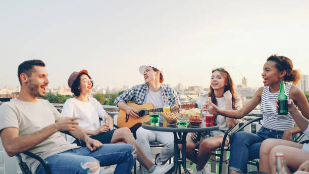 istock Creative guy is playing the guitar and his friends multi-ethnic group are singing holding bottles with beer and soft drinks sitting in circle at table on roof. 1436602142