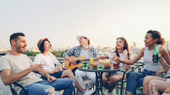 Creative guy in casual clothes is playing the guitar and his friends multi-ethnic group are singing holding bottles with beer and soft drinks sitting in circle at table on roof.