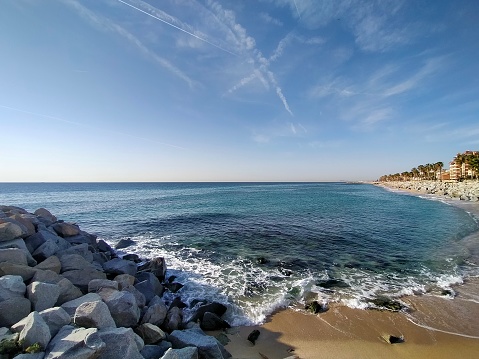 Escena de la playa de Vilassar de Mar con las rocas del rompeolas, el mar rompiendo en la arena con olas suaves y el cielo azul con nubes suaves y senderos de aviones. photo