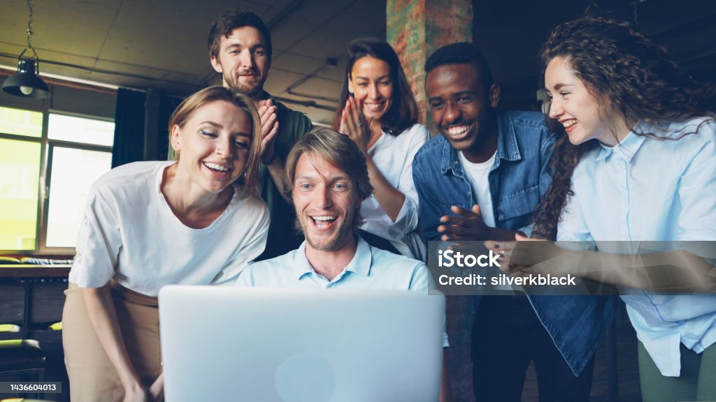 Excited young man is looking at laptop screen, rejoicing and expressing happiness, his colleagues are congratulating him on success clapping hands Excited young man is looking at laptop screen, rejoicing and expressing happiness, his happy colleagues are congratulating him on success, clapping hands People Stock Photo