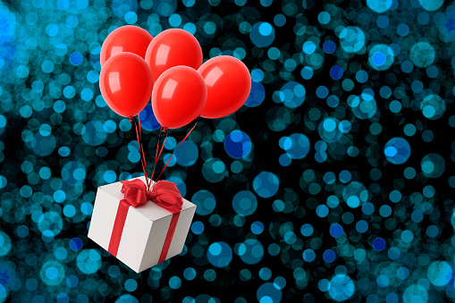 Christmas gift box suspended from red balloons and floating in mid-air, against blue defocused illumination.
