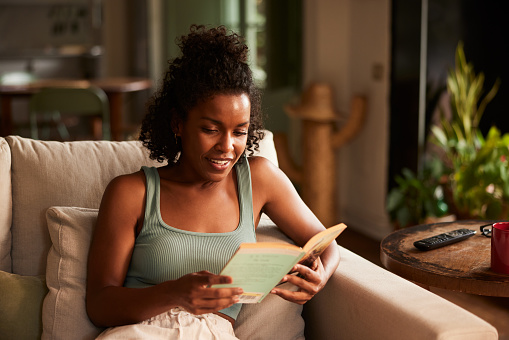 Sitting back on her living room sofa this attractive dark-skinned Latin American  woman is laughing to herself as she reads a funny story. Copy space in blurred background.