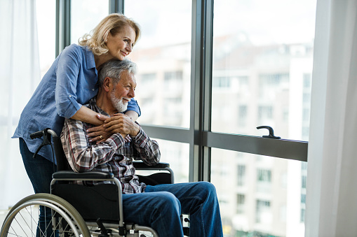 Senior woman and her husband in a wheelchair looking through window from their home. Copy space.