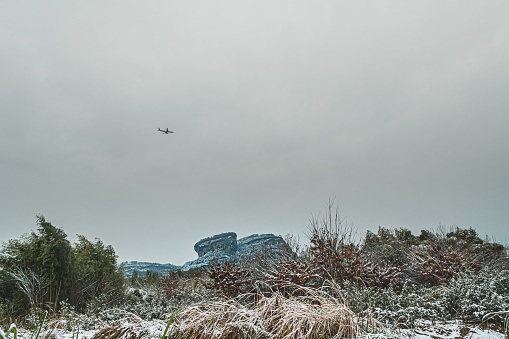 A scenic view of airplane flying in cloudy sky over rocky snow-covered hills and valleys on winter day