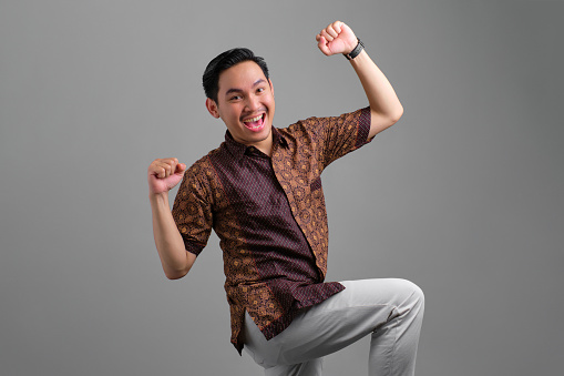 Excited young Asian man wearing batik shirt celebrating victory and raised fists isolated on grey background