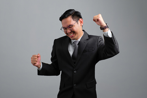 Excited young businessman in formal suit standing doing winner gesture clenching fists isolated on grey background