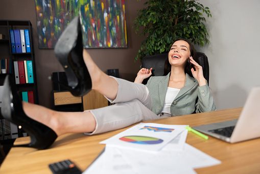 Beautiful delighted woman holding high heels shoes on desk near digital devices computer laptop papers having break after hard work day talking by cellphone smartphone discussing projects.