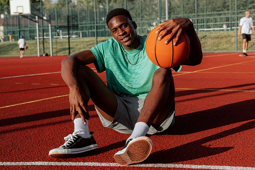 African american basketball player sitting on basketball court relaxing resting after exhausting training showing skills to friends comapny delighted man having break on background guys playing.