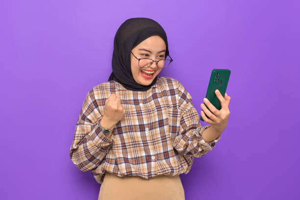 istock Excited young Asian woman in plaid shirt using a mobile phone, doing winner gesture, getting great news isolated on purple background 1436647970