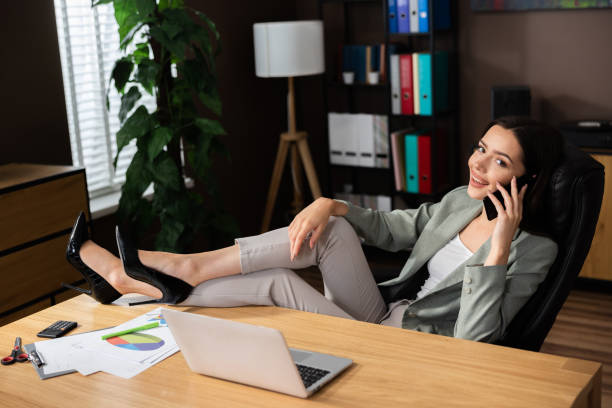 istock Working process conceot from home online. Beautiful delighted woman sitting in comfortable office chair holding legs on desk talking by cellphone smartphone. 1436648188