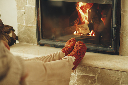 Heating house in winter with wood burning stove. Woman in cozy wool socks warming up feet at fireplace in rustic room. Young stylish female sitting at fireplace in farmhouse