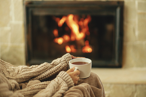 Hands in cozy sweater holding cup of warm tea on background of burning fireplace close up, autumn hygge. Heating house with wood burning stove. Relaxing and warming up at rustic fireplace