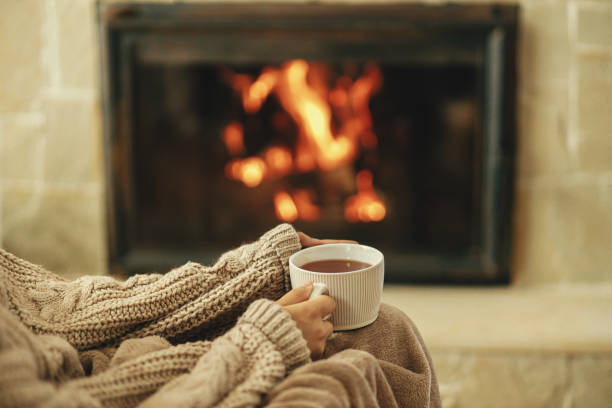 Hands in cozy sweater holding cup of warm tea on background of burning fireplace close up, autumn hygge. Heating house with wood burning stove. Relaxing and warming up at rustic fireplace stock photo