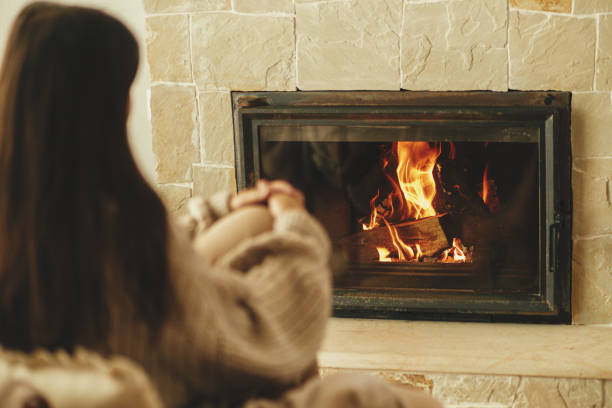 Chauffage de la maison en hiver avec poêle à bois. Femme élégante en pull confortable se réchauffant sur une chaise à la cheminée dans une chambre rustique. Jeune femme assise et se relaxant devant une cheminée dans une ferme - Photo