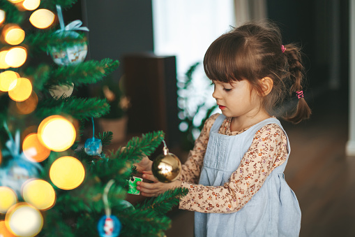 Portrait of a cute little girl hanging ornaments on a Christmas tree with lights and balls. Kid decorating Christmas tree.