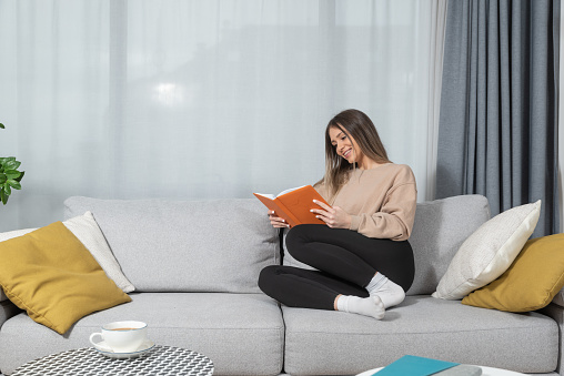 People and leisure concept - young woman reading book on sofa at home enjoying her free time from study and working in her home.