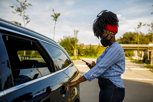 Copy space shot of young Black woman opening the back door and entering a taxi after ordering a ride via mobile app on smart phone. She is wearing a protective face mask.