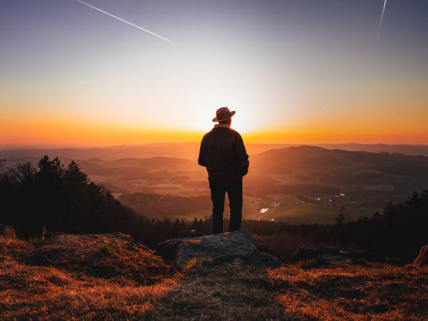 istock Man with a hat standing on a mountain peak and enjoying a beautiful landscape during sunset 1436677156