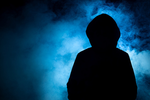 A closeup of a man with a dark black top with a blue smoke background, technology, and cybercriminal concepts