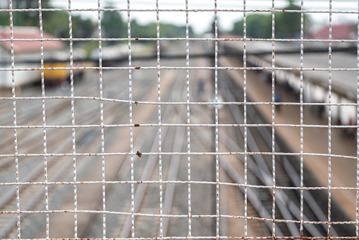 Metal wire fence wall with train station and railway track as blurred background. Close-up and selective focus.