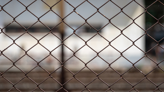 Metal wire fence wall with train station and railway track as blurred background. Close-up and selective focus.