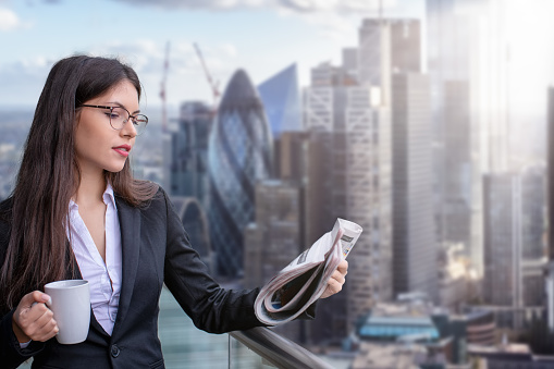 A attractive businesswoman in corporate outfit reading morning newspaper and holding a coffee cup in front of the City of London skyline