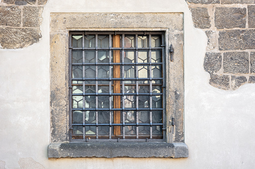 Rectangular window with a metal grate against the background of a peeling plastered gray stone wall. From the Window of the World series.