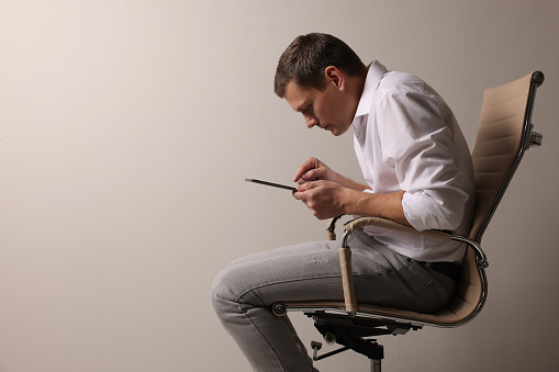 Man with bad posture using tablet while sitting on chair against grey background. Space for text