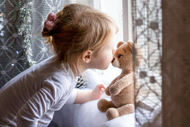 bambina carina che sorride baciando il suo orsacchiotto giocattolo vicino alla finestra di luce. concetto di infanzia, amore, gentilezza, amicizia - 4 of a kind foto e immagini stock