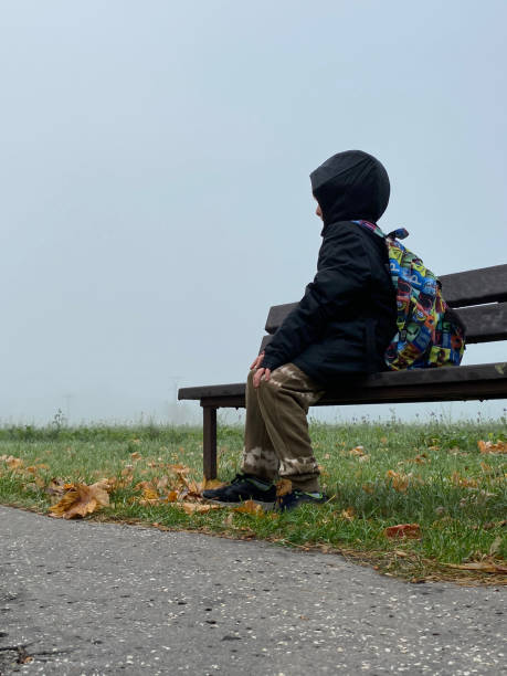 istock dramatic scene of a kid with a backpack alone sitting on a park bench, in a cold and foggy winter day waiting for his mum or father. Boy is lost in the dark 1436716082