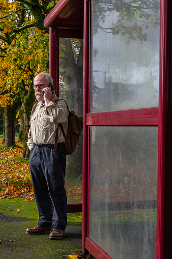 Retired man standing alone at a rural bus stop while he is talking on a mobile phone in Scotland